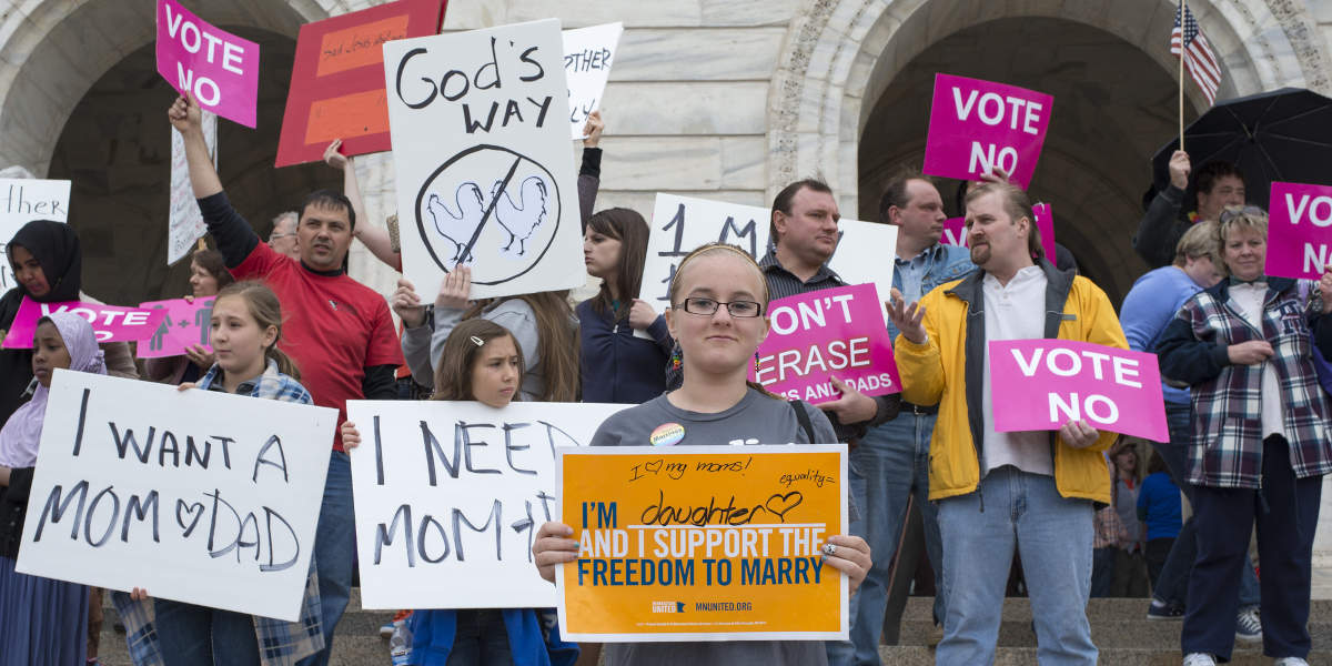 On the steps of a government building, protestors hold signs for (a young girl smiling in the foreground) and against (a mixed-age crowd in the background) same-sex marriage