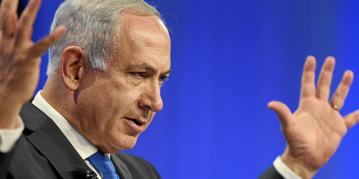 A close up image of a white man in a suit, Netanyahu, holding arms aloft against a blue background