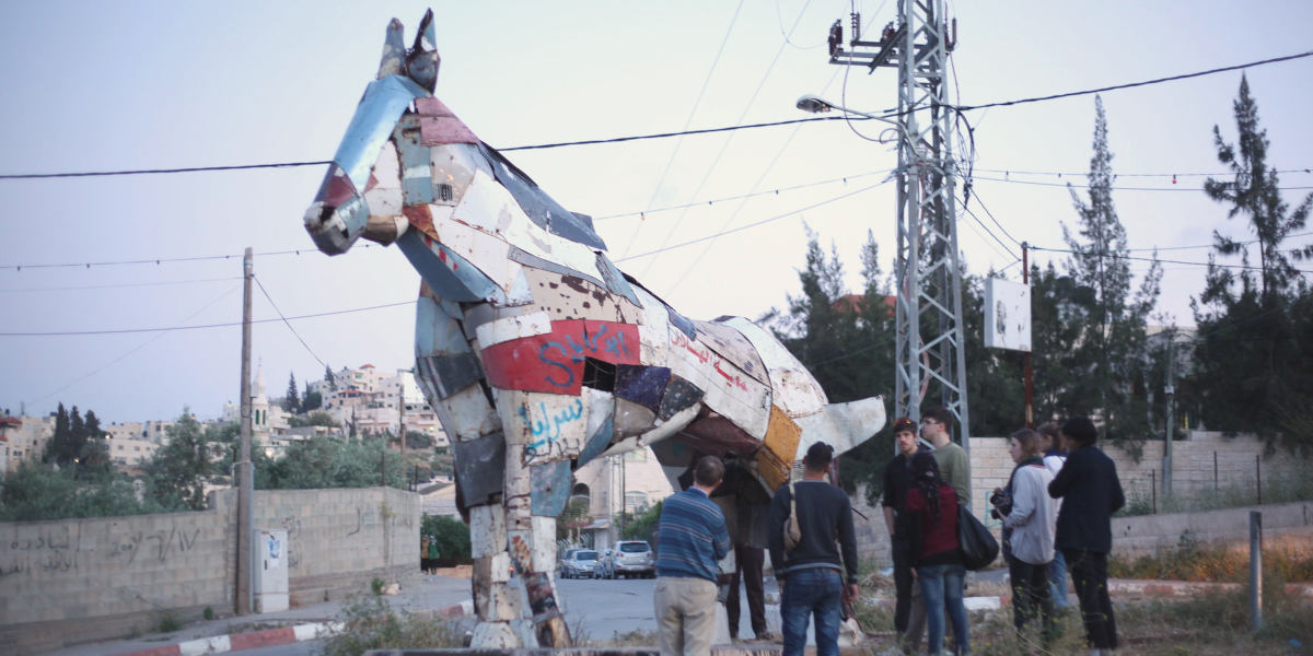 A group of people look up at a statue of a horse made from scrap metal, with roads and buildings in the background. The statue is in Jenin, Palestine