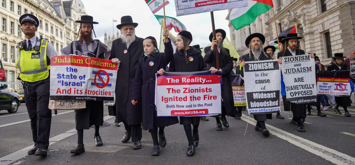 A group of Orthodox Jewish men and children in traditional Hasidic clothing holding placards reading 'Judaism condemns the state of Israel and its attrocities'