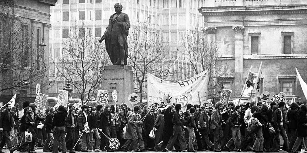 A black and white photo of Rock against Racism protestors, holding banners and placards, marching near the National Gallery in London in 1978
