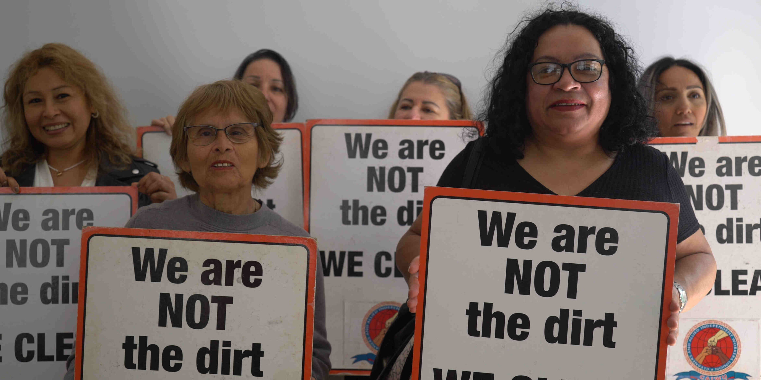 Six women stand together holding signs that say 'We are NOT the dirt we clean'.