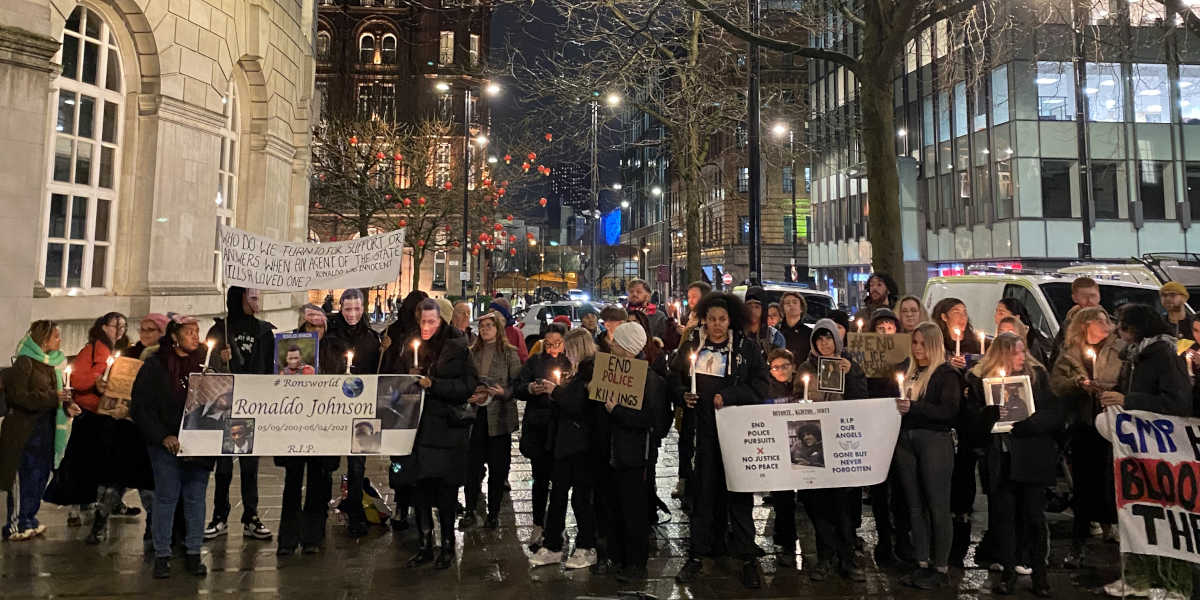 A gathering of 50 people holding candles, banners and statements of support stand together at night outside a large white stone building