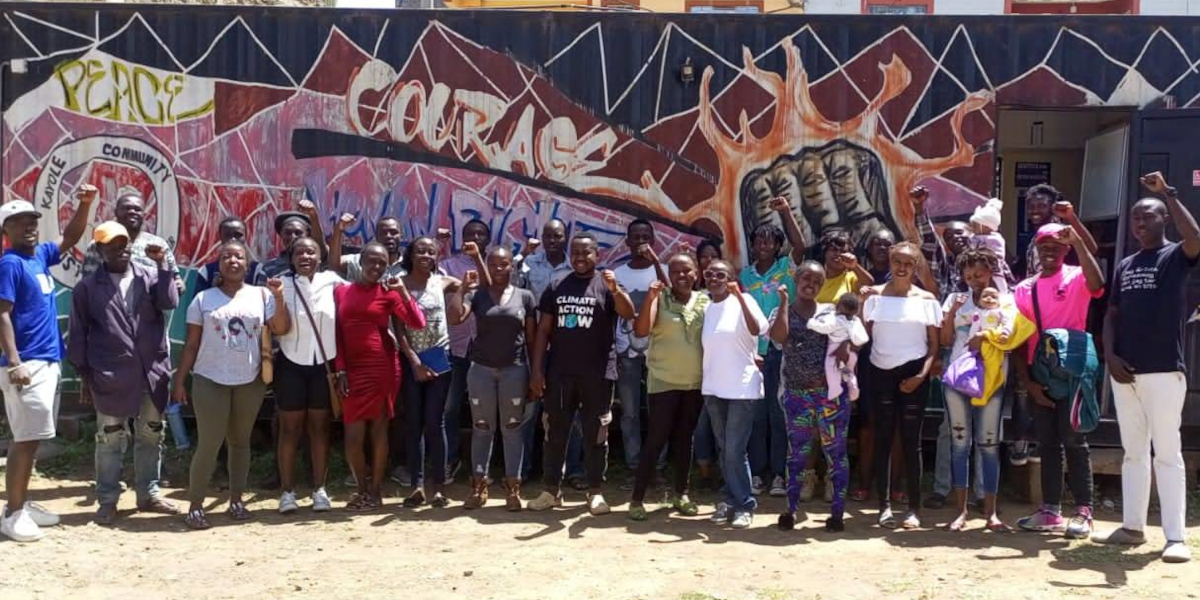 Members of the Kayole Social Justice Centre stand in front of a wall of grafiti with their fists raised. The wall says 'peace' and 'courage'