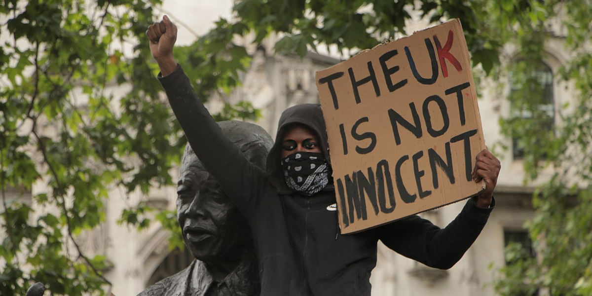 A masked protestor stands in front of a statue of Nelson Mandela with a raised fist holding a sign saying 'The UK is not innocent'