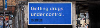A blue Transform Drug Policy Foundation billboard under a train bridge reading 'Getting Drugs Under Control.'