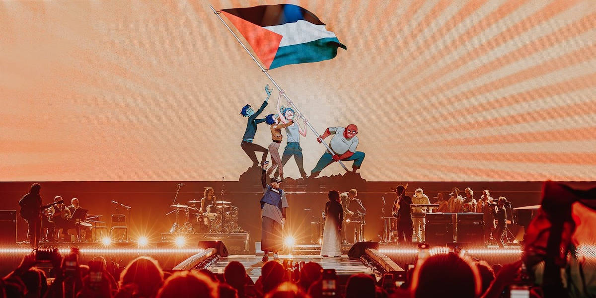 A photograph of musicians on stage from the crowd, with artwork showing a Palestine flag being hoisted into the air