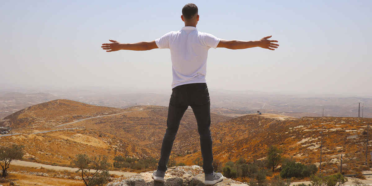 A person in jeans and a white shirt stands arms outstretched looking over a valley