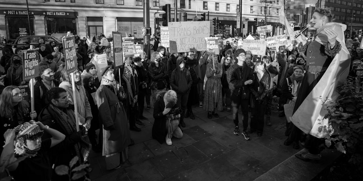 A black and white photo of a crowd of people stood in a semi-circle listening to a speaker, whilst holding placards reading 'trans rights now' and 'trans bodies are not a political playground'