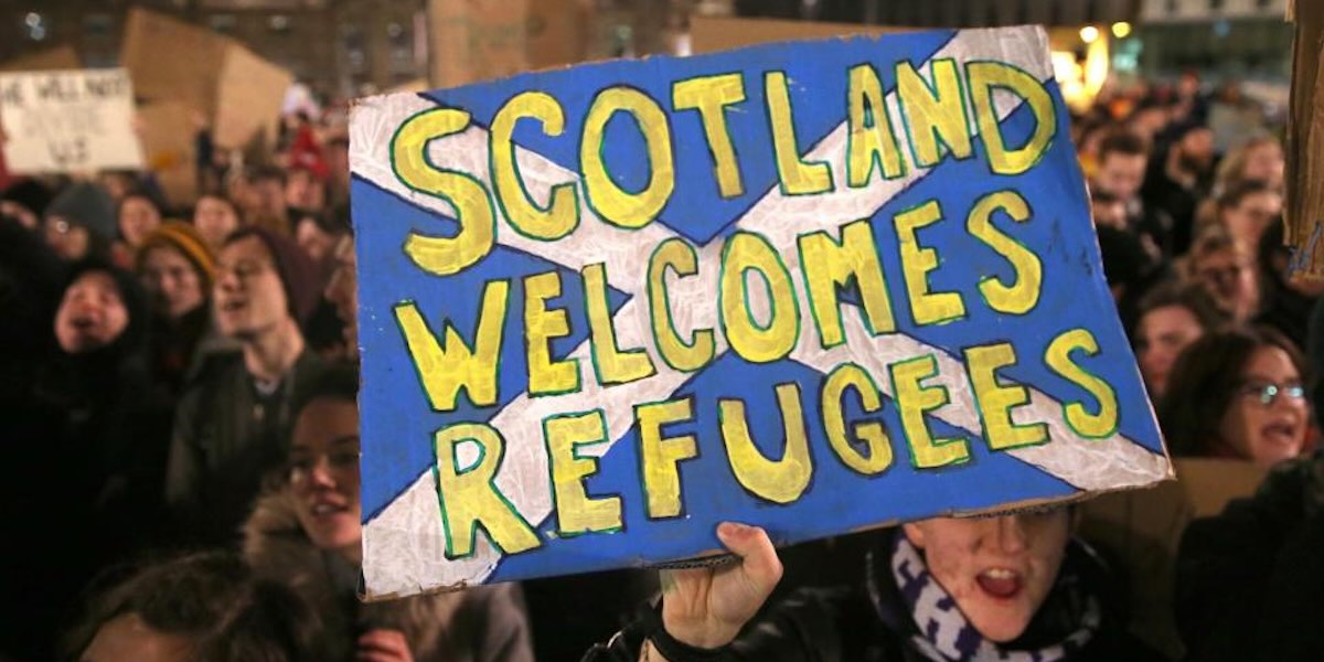A close up of a hand painted sign that reads 'Scotland Welcomes Refugees' against the national flag
