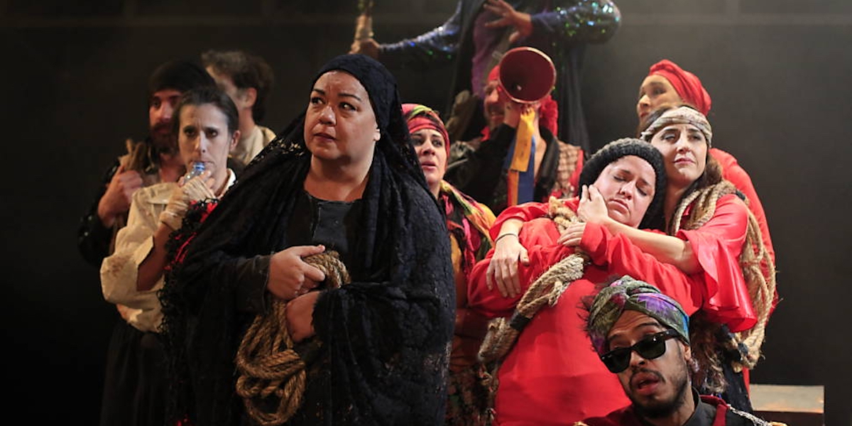A theatrical scene sees a woman in black surrounded by a group wearing red, with white faces