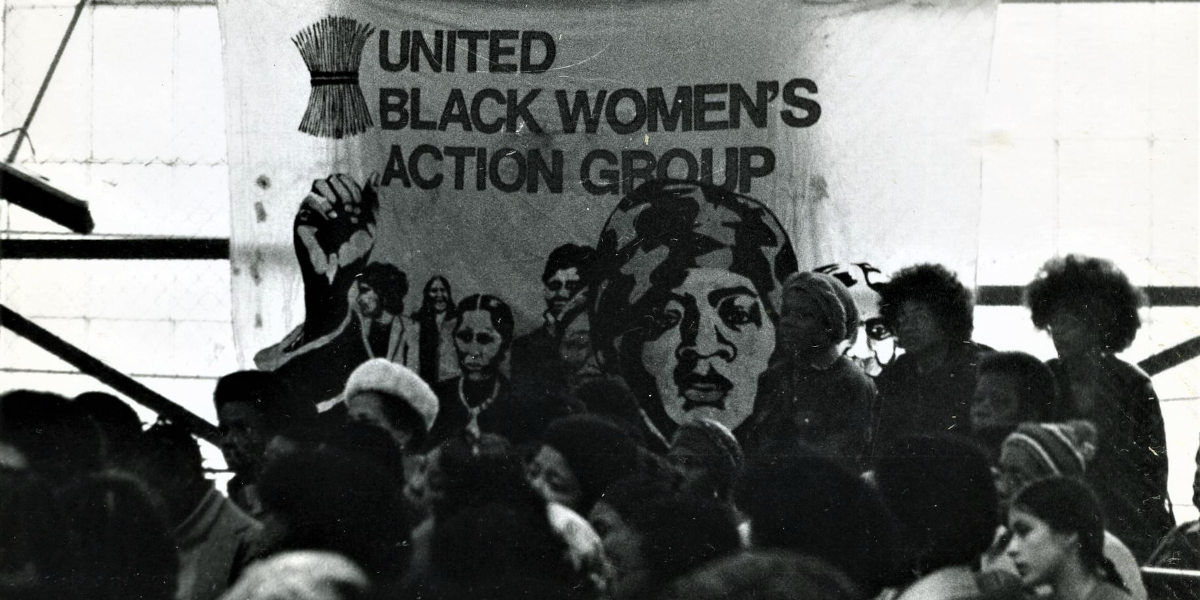 A black and white photo of a crowd with a banner in the background reading 'United Black Women's Action Group'