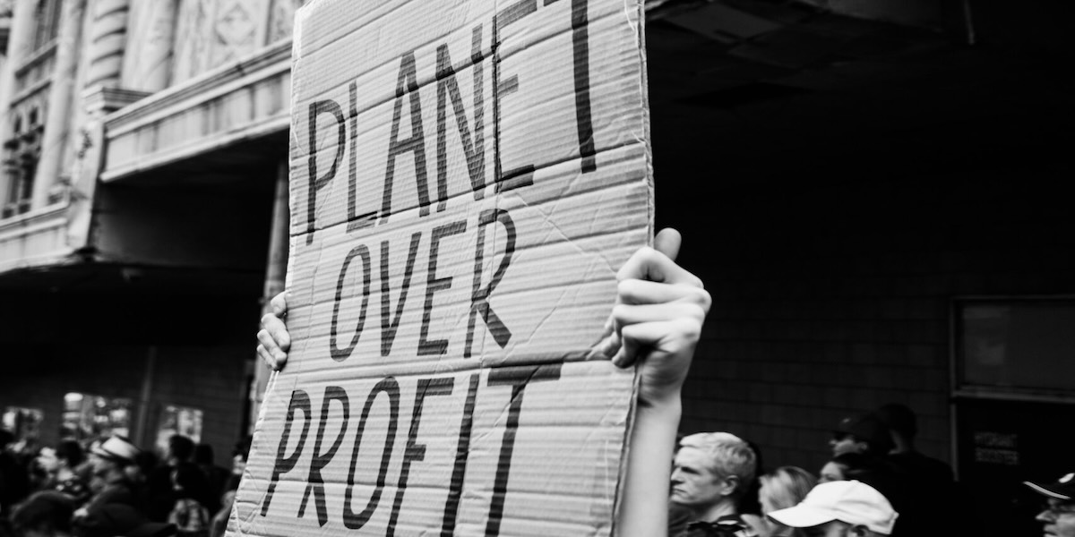 A black and white photo of hands holding aloft a cardboard sign that reads: 'planet over profit'