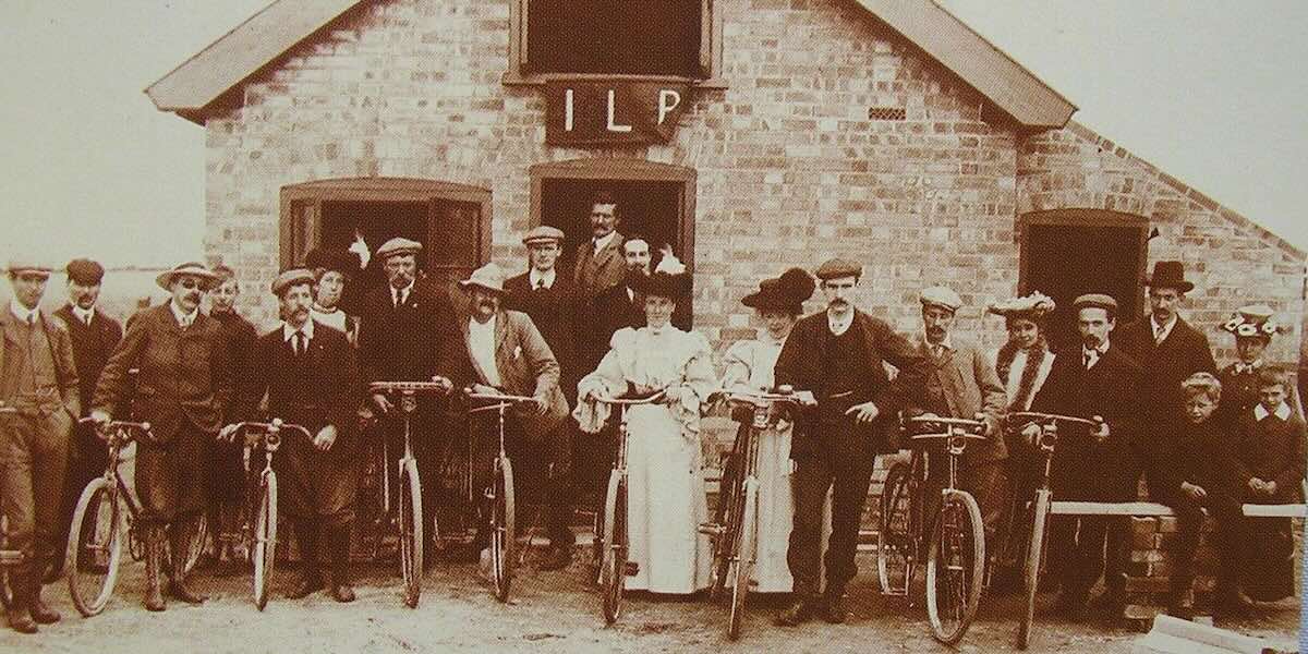 A sepia photograph shows men and women with bikes in front of a building with an ILP banner