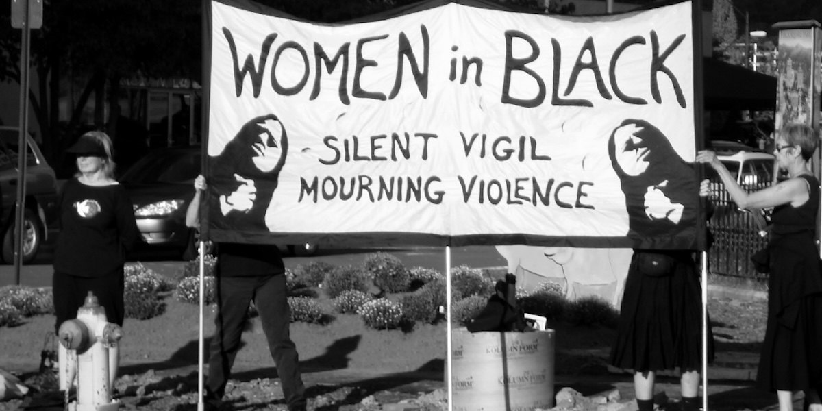 A black and white photograph of a vigil held by Women in Black, with a group of women holding a banner reading 'women in black silent vigil mourning violence'